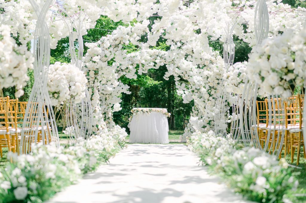 An elegant outdoor wedding venue setup with white floral arches and chairs.
